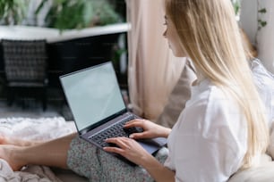 Bloguera adulta joven sentada en una cama cómoda en pijama, crea contenido para su blog en línea en una computadora portátil. Mujer mirando el monitor del espacio de copia y pasando la mañana en casa en un dormitorio acogedor y luminoso