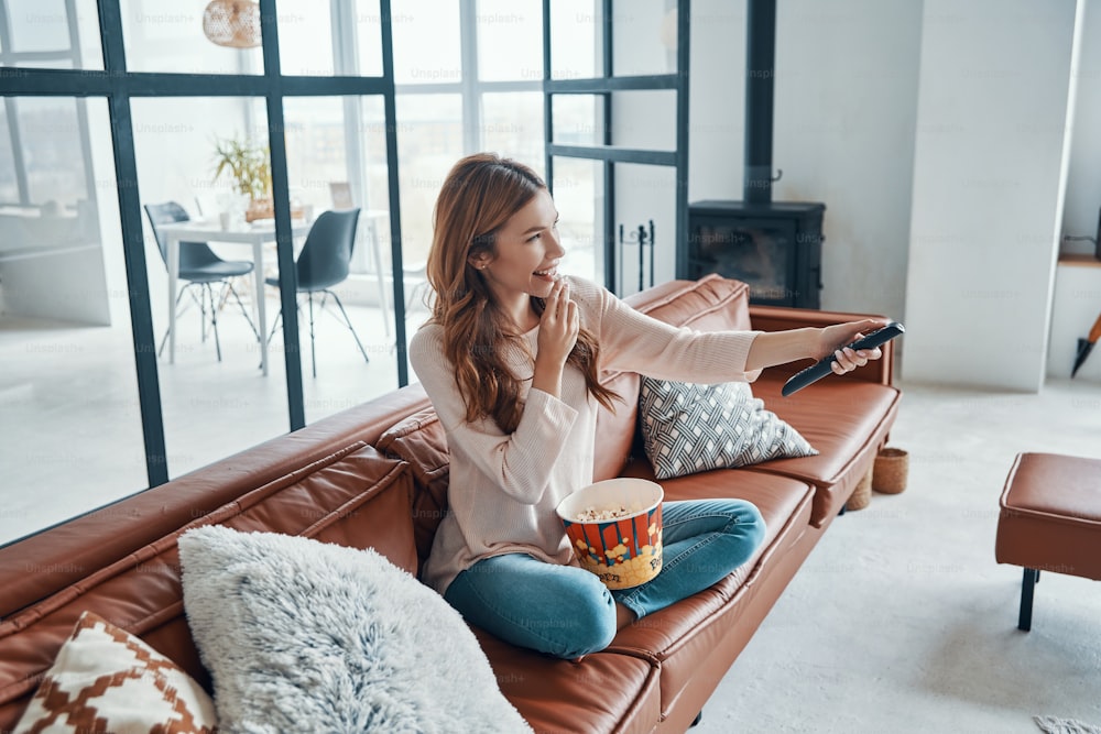 Beautiful young woman watching television and eating popcorn while sitting on the sofa at home