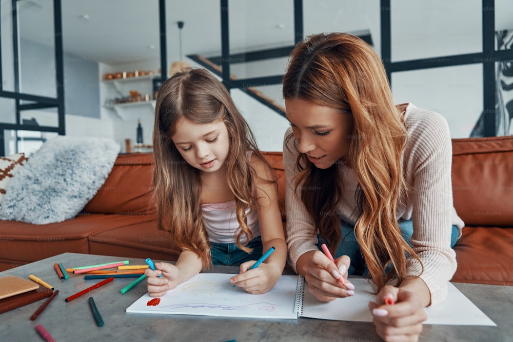 Jovem bela mãe e sua filha pequena desenhando em caderno de esboços enquanto passam o tempo em casa