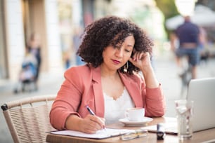 Woman working on paperwork. Business woman sitting in café using laptop and writing on document.