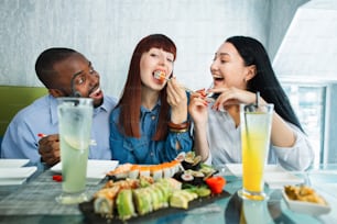Sushi concept, seafood, asian meal. Three young multiethnic friends, Caucasian and Asian girls, African man, sitting in restaurant, spending funny time with plate of delicious sushi