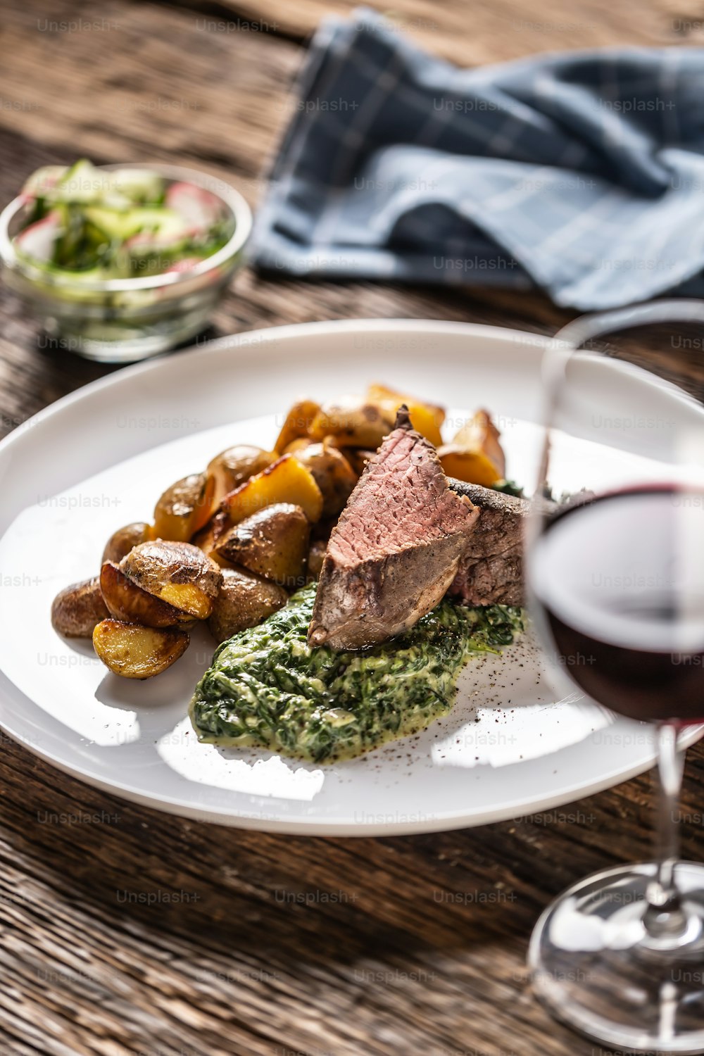 Red wine as a typical baverage to a red meat sous vide steak with roasted potatoes, spinach dip and fresh salad served on a white porcelain plate on the wooden desk.