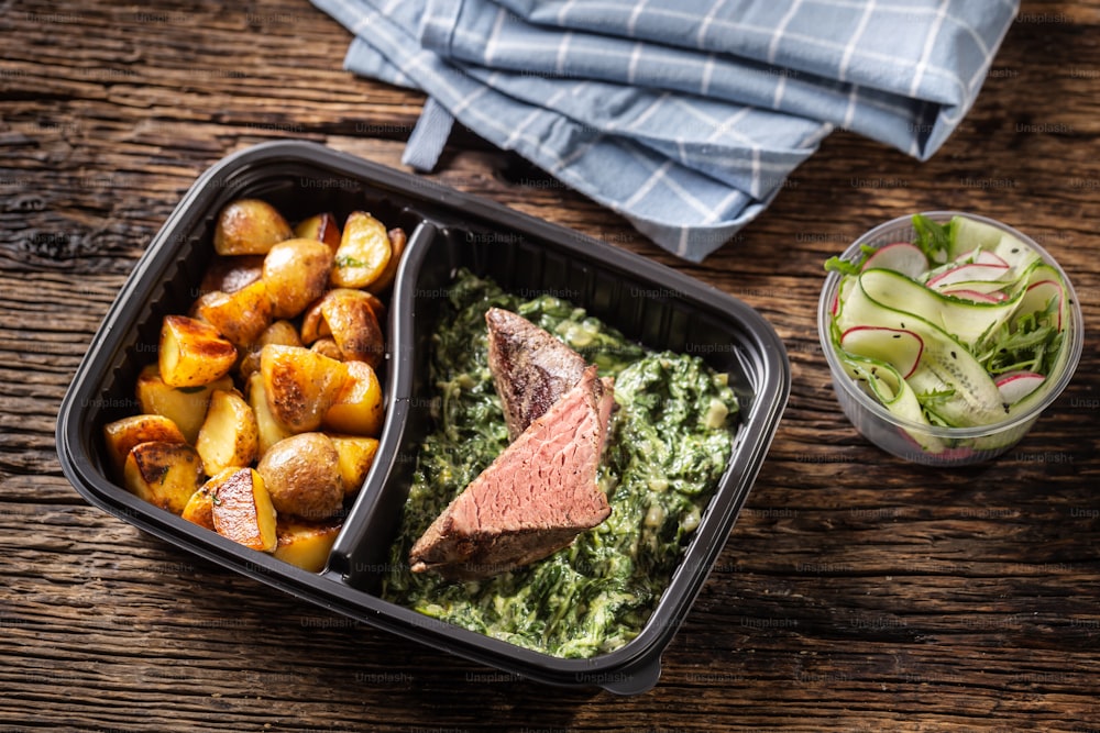 Delicious red meat sous vide steak packed in portable take away plastic box and served with crunchy roasted potatoes and savory spinach dip and fresh vegetable salad.