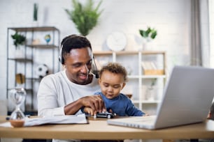 Casual african businessman in headset having working meeting on laptop while nursing his baby son at home. Concept of technology and freelance.