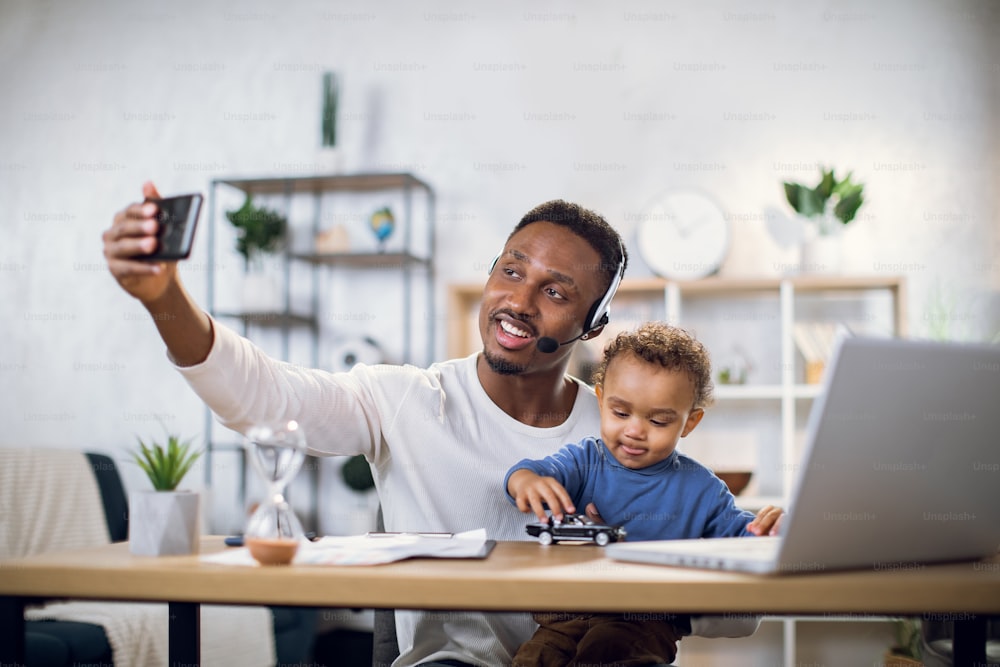 Smiling african man taking selfie on smartphone while sitting at table with son on knees. Young father using modern laptop for remote work from home.