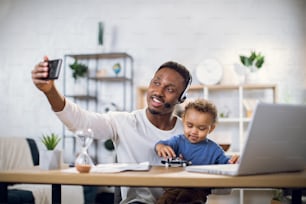 Smiling african man taking selfie on smartphone while sitting at table with son on knees. Young father using modern laptop for remote work from home.