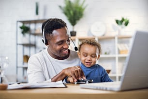 헤드셋을 쓴 아프리카계 미국인 아버지는 귀여운 아들과 놀고 현대 노트북에서 온라인 회의를 하고 있다. 젊은 남자는 아기와 함께 집에 앉아 거리를 두고 일하고 있다.