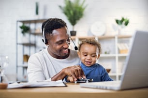 Afro american father in headset playing with cute son and conducting online meeting on modern laptop. Young man sitting at home with baby boy and working on distance.