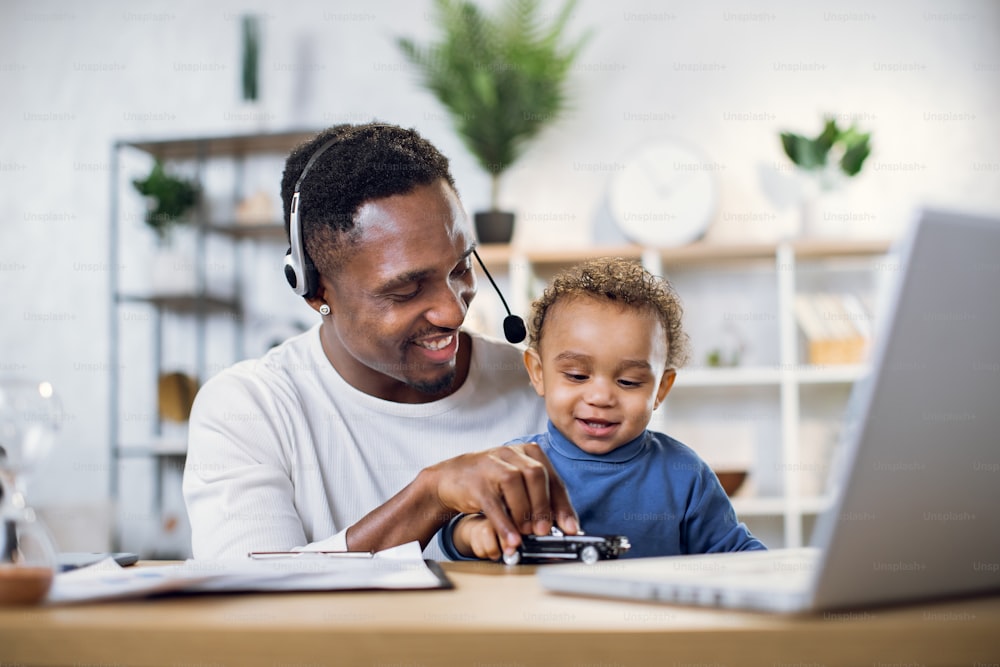 Afro american father in headset playing with cute son and conducting online meeting on modern laptop. Young man sitting at home with baby boy and working on distance.