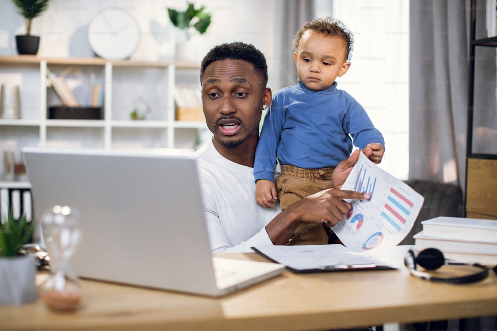 Afro american businessman having online meeting on modern laptop while carrying his baby son on hands. Young father working remotely from home.