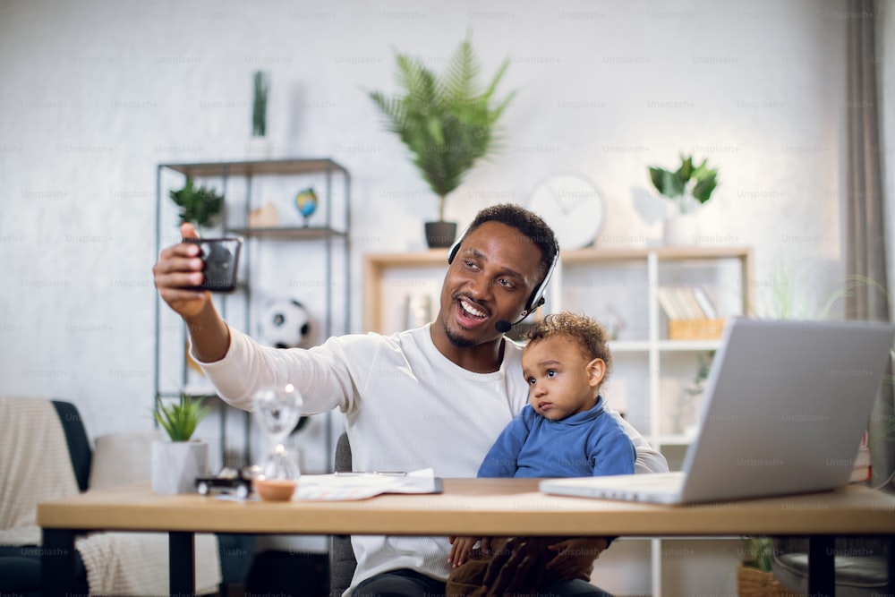 African male freelancer in headset carrying baby and using mobile for taking selfie. Young father with son working remotely from home office.