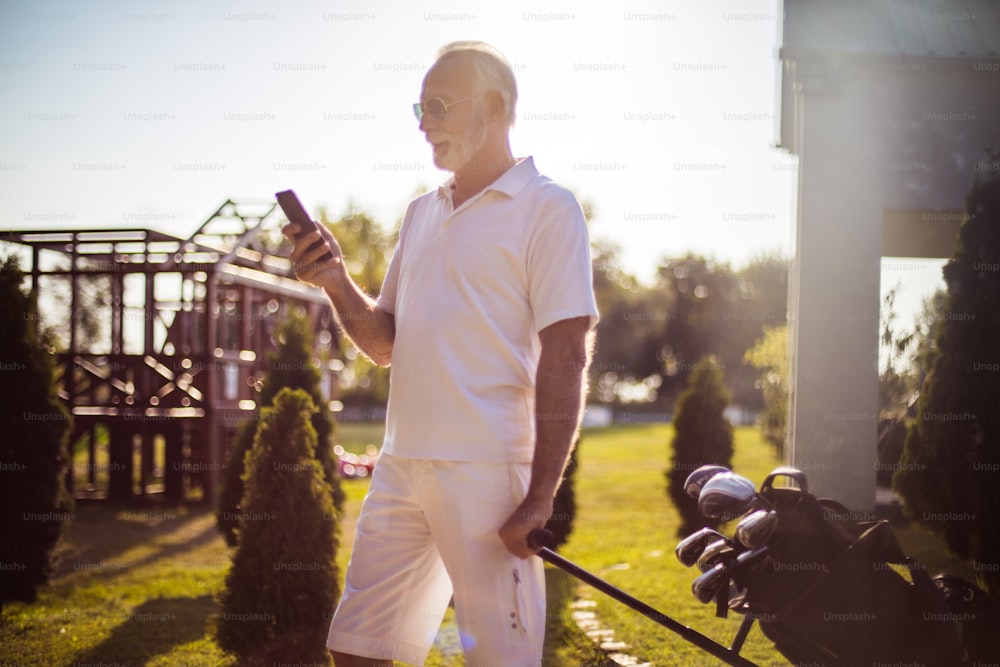 Senior man walking with golf bag and using phone.