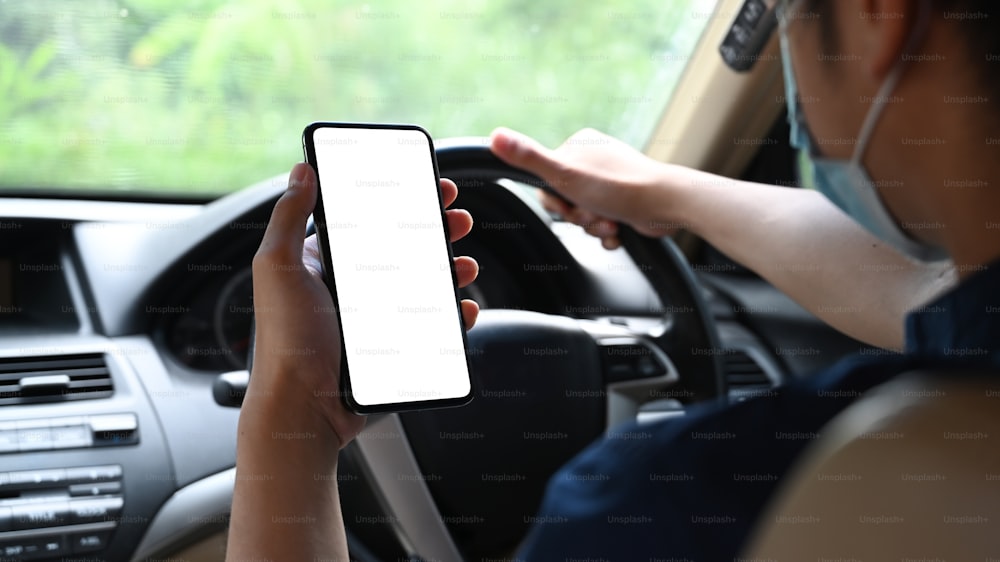 Foto recortada de un joven con máscara protectora y un teléfono inteligente mientras conduce un automóvil.