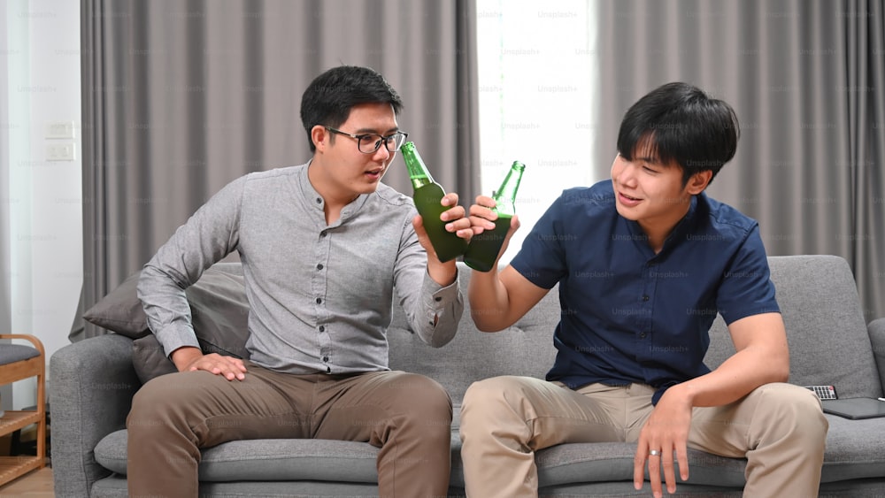 Happy two men drinking beer while sitting on sofa.