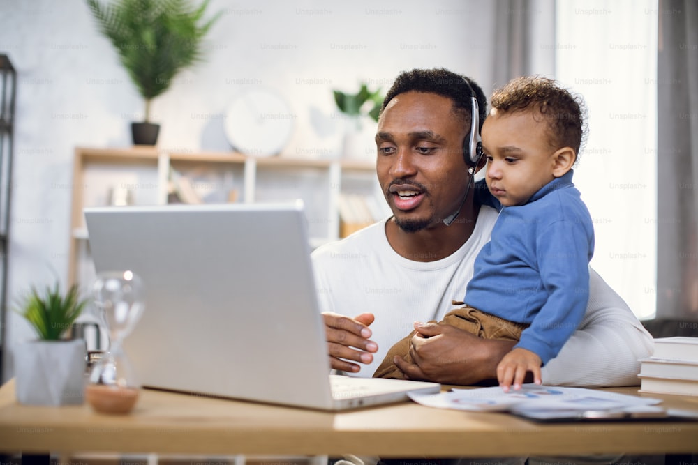 Afro american father in headset using modern laptop for video conference while holding baby boy on hands. Male freelancer with so trying to work on distance.