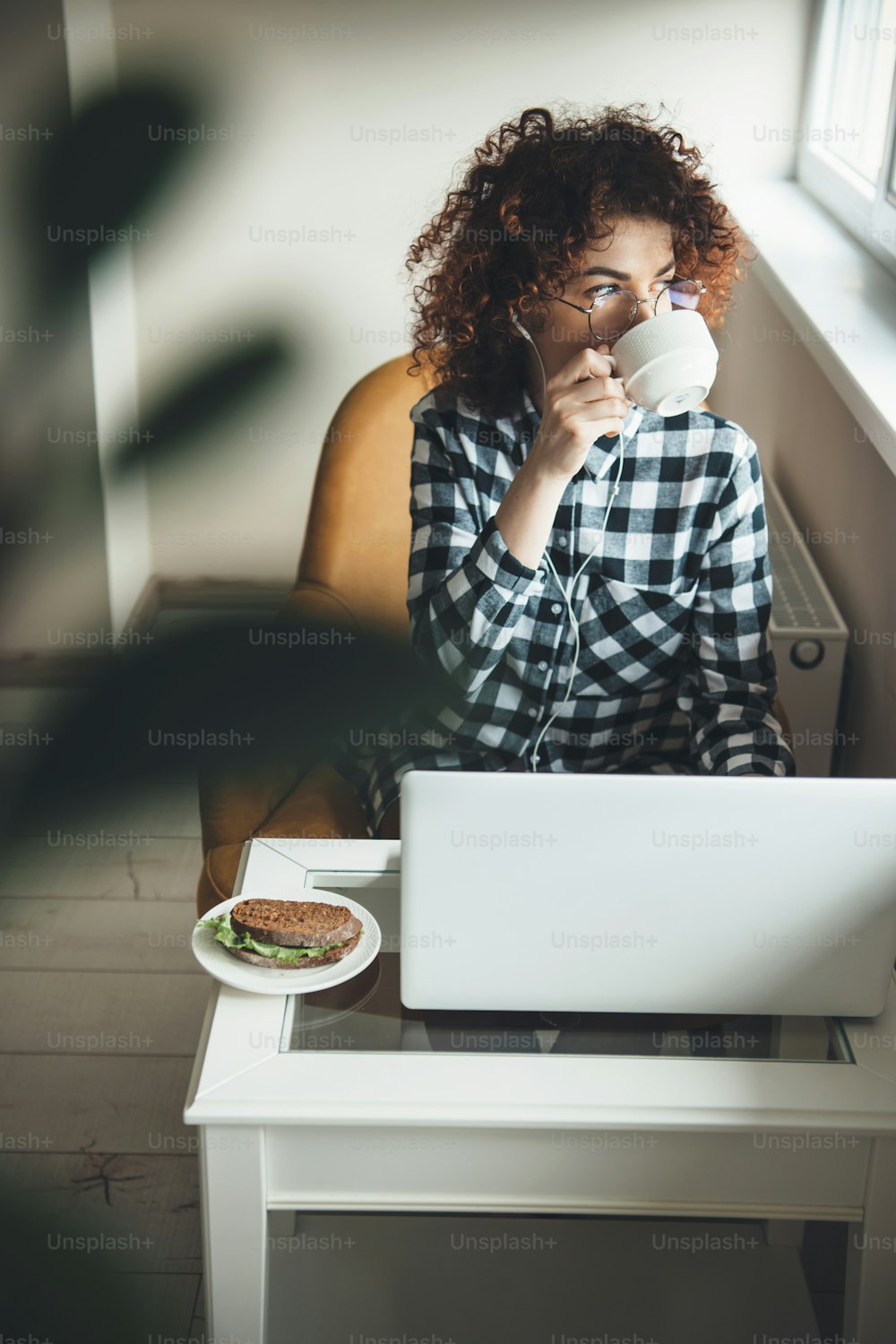 Young curly businesswoman drinking a coffee with sandwich while working at the computer