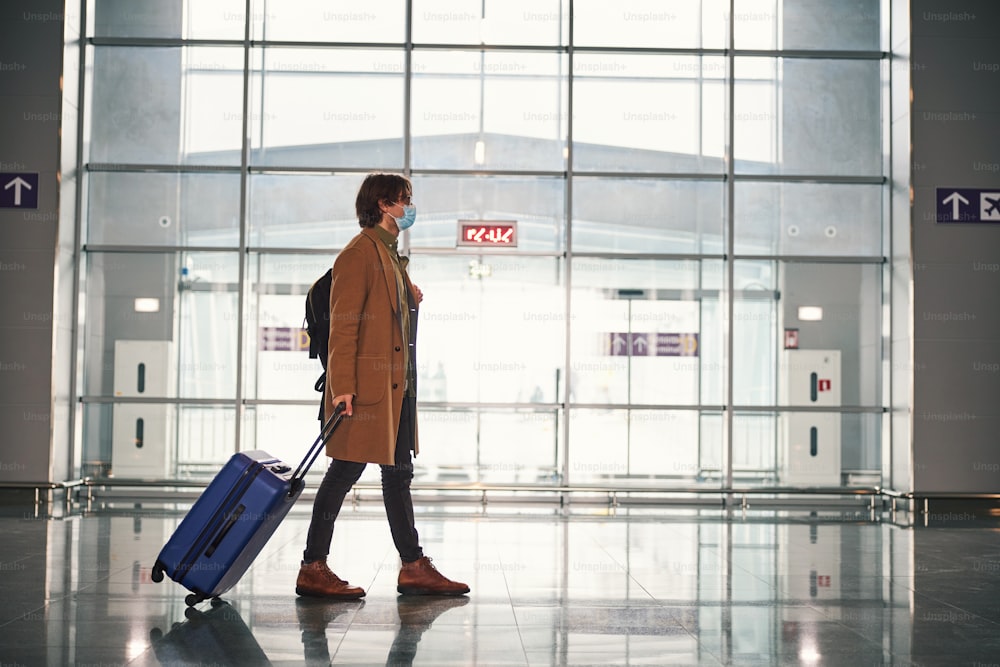 Persona di sesso maschile a tutta lunghezza con borsa trolley bagagli che indossa una maschera protettiva per il viso mentre cammina lungo il terminal dell'aeroporto