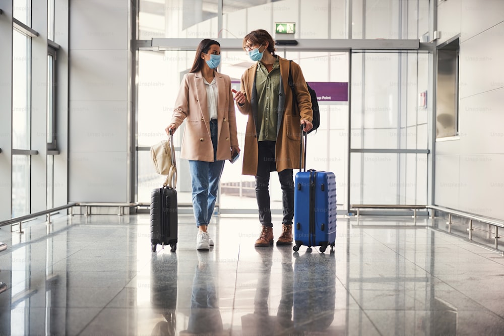 Uomo e donna alla moda con valigie da viaggio che indossano maschere protettive per il viso mentre chiacchierano e aspettano il volo in aeroporto
