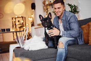 Uomo sorridente in jeans seduto sul divano di casa con il suo simpatico cane.
