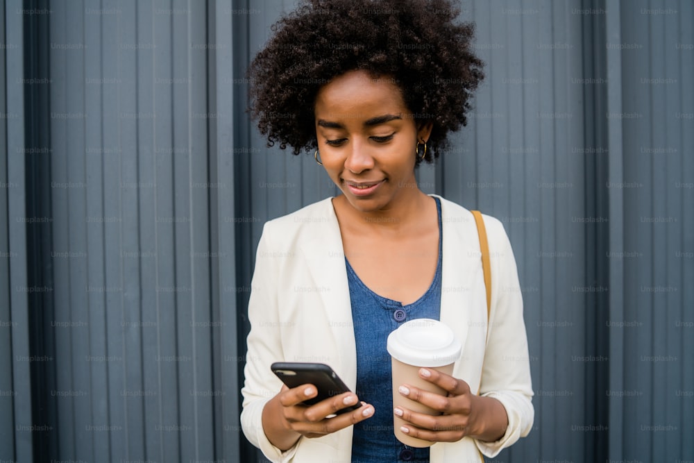 Retrato de una mujer de negocios afro usando su teléfono móvil y sosteniendo una taza de café mientras está de pie al aire libre en la calle. Concepto empresarial y urbano.