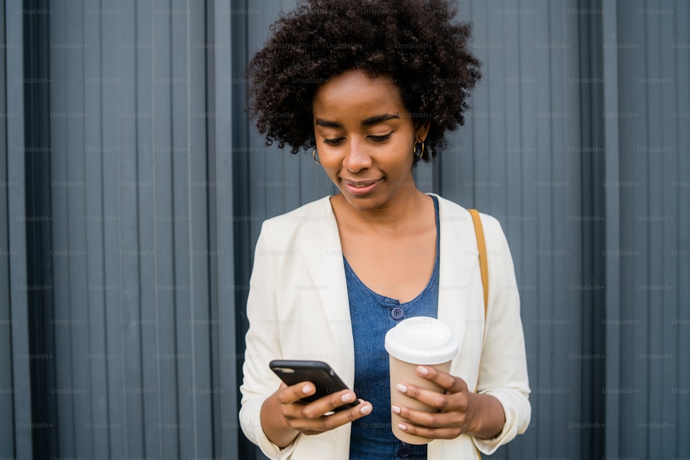 Ritratto di donna d'affari afro che usa il suo telefono cellulare e tiene una tazza di caffè mentre si trova all'aperto in strada. Concetto di business e urbano.