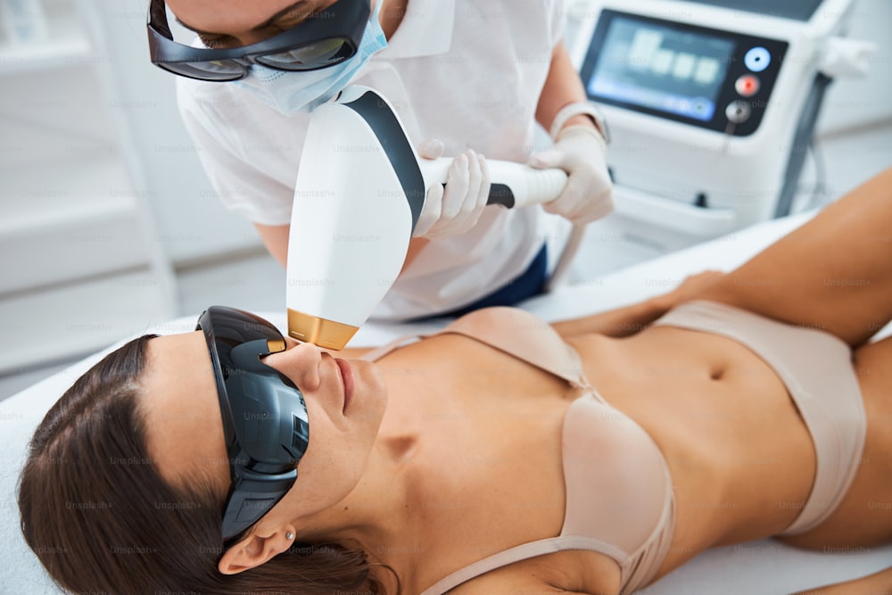Focused dermatologist in a mask and safety goggles pointing the laser beam at the woman face