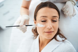 Vista superior de uma mulher deitada com os olhos fechados durante um procedimento de beleza conduzido por um cosmético