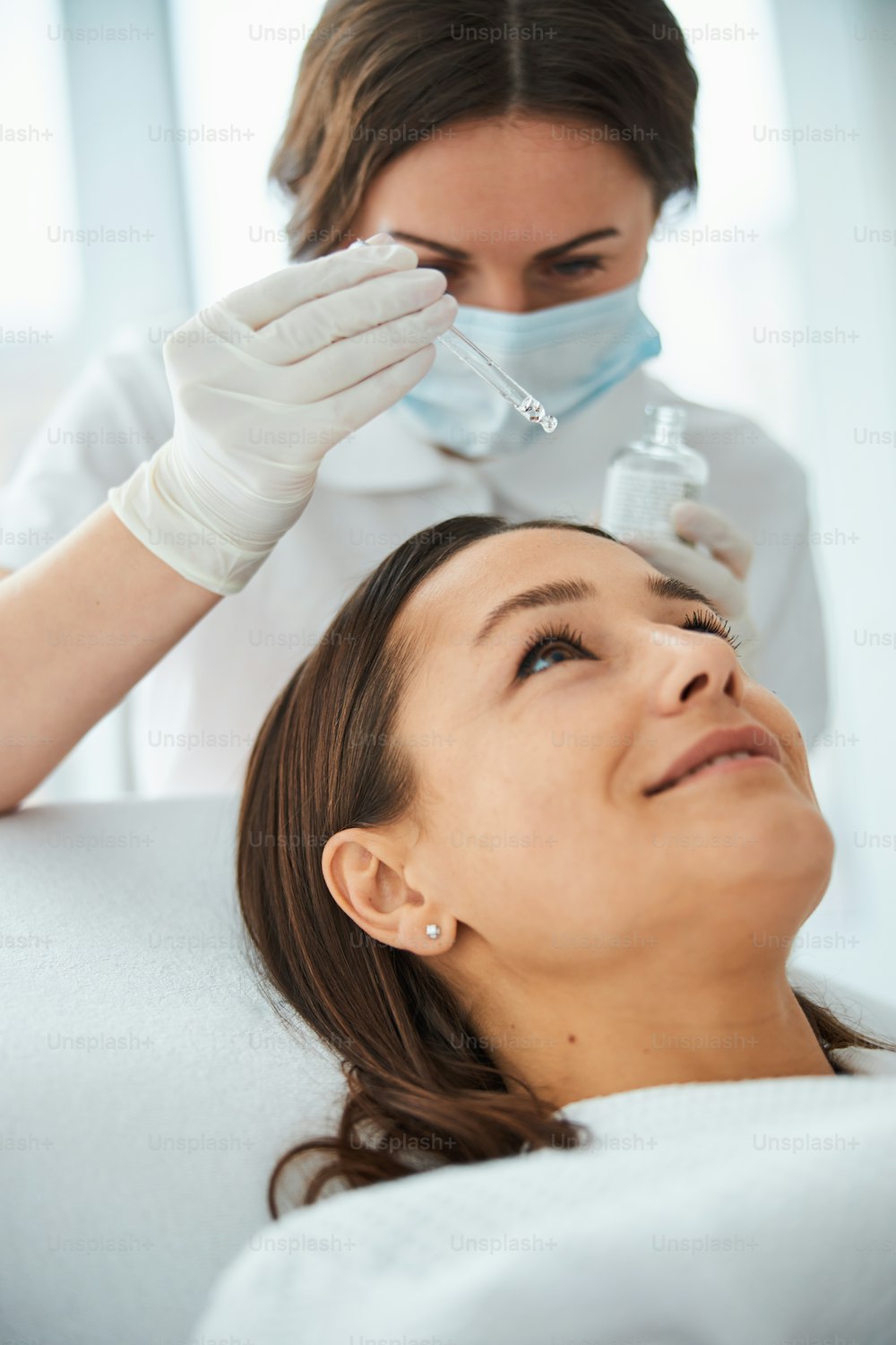 Medico concentrato in maschera facciale e guanti che applica il siero sulla pelle femminile utilizzando una pipetta