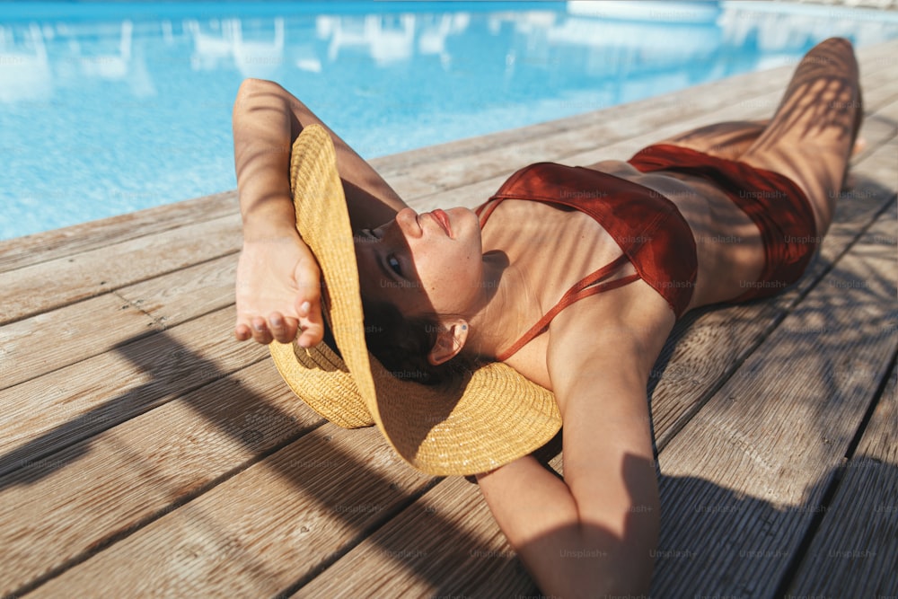 Beautiful woman in hat relaxing on wooden pier under palm leaves at pool, enjoying summer vacation. Slim young female lying and tanning at swimming pool at tropical resort. Travel
