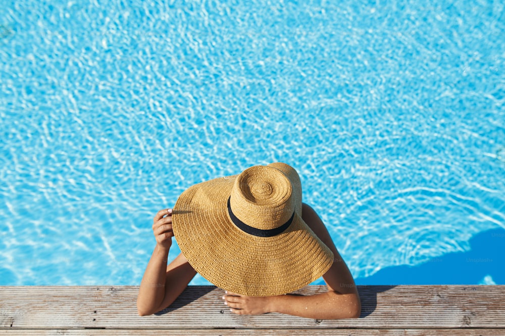 Beautiful tan woman in hat relaxing in pool water at wooden pier, enjoying summer vacation, top view. Slim young female sunbathing at stylish swimming pool edge. Space for text. Travel and Holidays