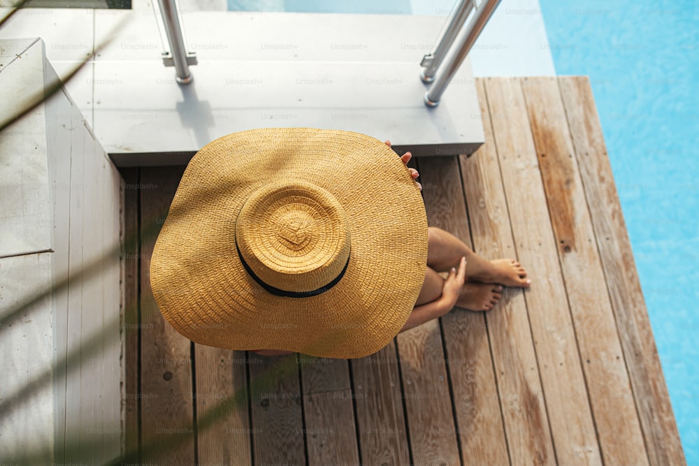 Beautiful woman in hat relaxing on wooden pier at blue pool enjoying summer vacation. Top view of slim young female sunbathing and tanning at swimming pool. Holidays and travel concept
