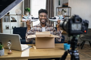 Smiling african man sitting at table with new sneakers in hands and recording video review on professional camera. Concept of blogging, advertising and modern technology.