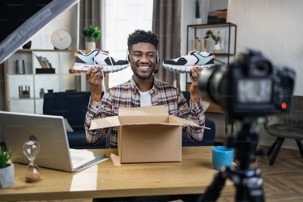Uomo africano sorridente seduto al tavolo con nuove scarpe da ginnastica in mano e registrazione video recensione su fotocamera professionale. Concetto di blogging, pubblicità e tecnologia moderna.