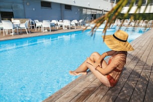 Beautiful woman in hat tanning on wooden pier at blue pool enjoying summer vacation. Slim young female relaxing at tropical resort and sunbathing at swimming pool. Travel and Holidays