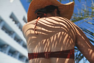 Shadow of palm leaves on back of beautiful woman in hat relaxing at pool enjoying summer vacation. Skin care and sunscreen concept. Slim young female tanning at swimming pool at tropical resort