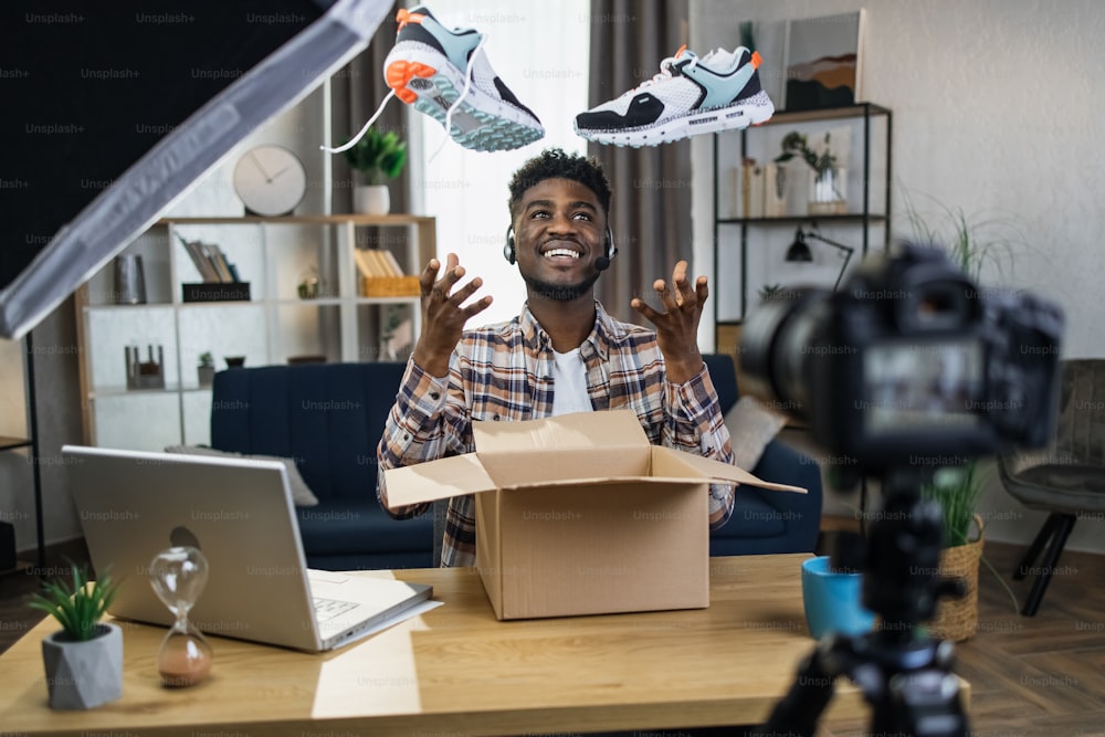 Fröhlicher Afro-Mann im Headset, der Videoinhalte für seine sozialen Netzwerke erstellt, während er zu Hause neue Turnschuhe auspackt. Glücklicher Blogger, der sein Lieblingshobby mit Hilfe moderner Gadgets genießt.
