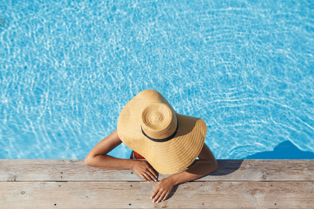 Godersi le vacanze estive. Donna elegante in cappello che si rilassa nell'acqua della piscina al molo di legno, vista dall'alto. Giovane femmina magra che prende il sole sul bordo della piscina. Viaggi e vacanze. Spazio per il testo