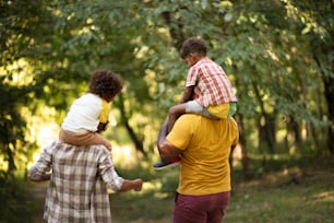 Família afro-americana caminhando pelo parque. Pais carregando crianças em carona.