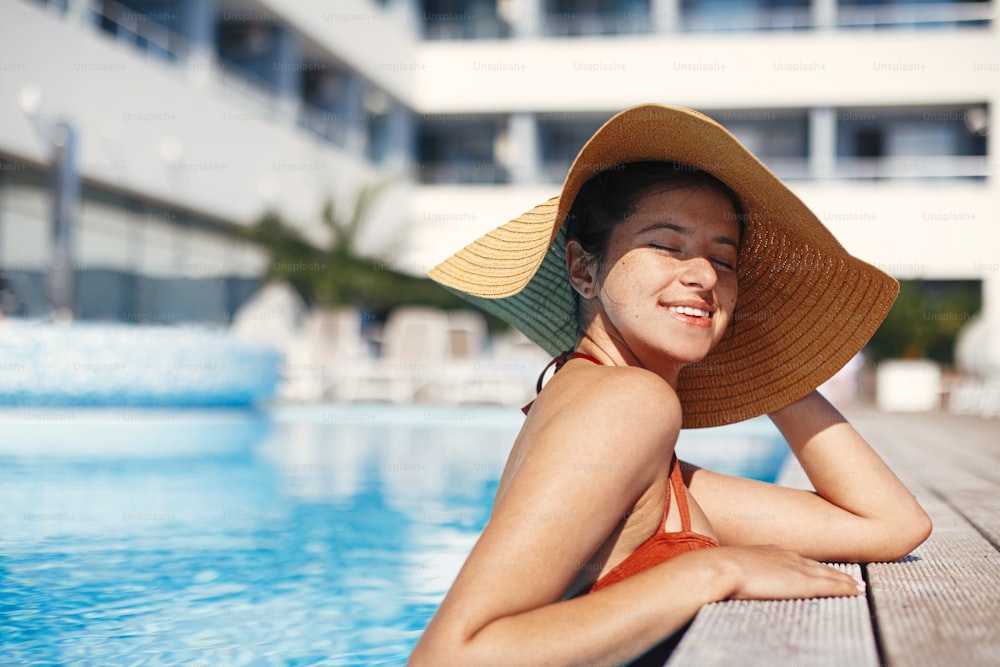Enjoying summer vacation. Beautiful happy woman in hat relaxing in pool water. Stylish slim young female sunbathing and resting at swimming pool edge at tropical resort. Holidays and travel