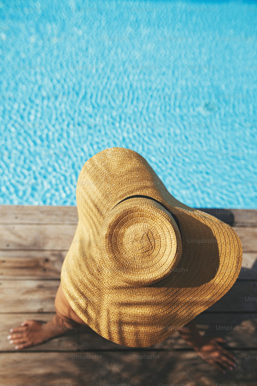 Enjoying summer vacation. Beautiful woman in hat relaxing on wooden pier under palm leaves shadow at edge of pool. Top view of slim young female sunbathing at swimming pool. Holidays and travel