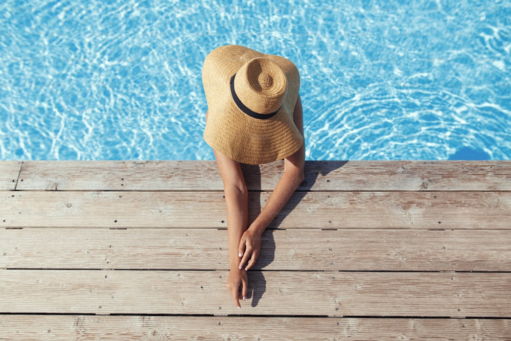 Enjoying summer vacation. Beautiful tan woman in hat relaxing in pool water at wooden pier, view above. Slim young female sunbathing at swimming pool edge at tropical resort. Travel and Holidays