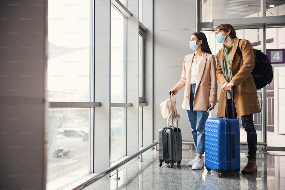 Giovane uomo e donna alla moda con valigie da viaggio che indossano maschere protettive per il viso mentre guardano fuori dal finestrino e aspettano il volo