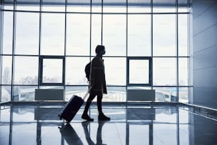 Passageiro do sexo masculino com saco de carrinho de bagagem usando máscara facial protetora enquanto caminha pelo terminal do aeroporto