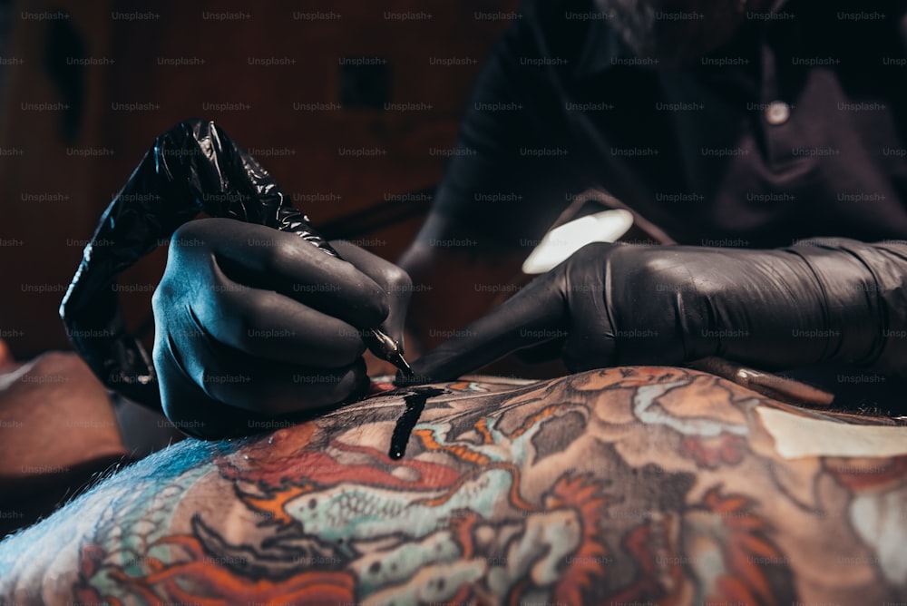 Tätowiererhände, die schwarze Schutzhandschuhe tragen und eine Maschine halten, während sie ein Bild auf einer Haut erstellen und die Tinte tropft