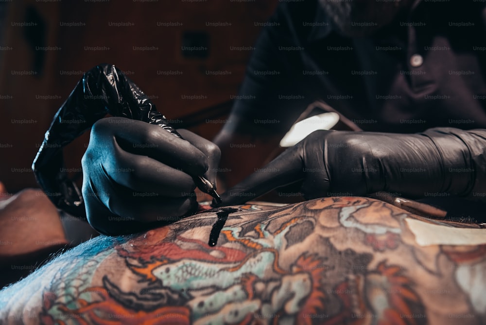Tattoo artist hands wearing black protective gloves and holding a machine while creating a picture on a skin and the ink is dripping