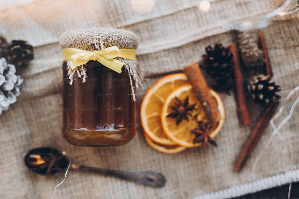 Honig im Glas mit Band und Löffel, Anisstern, Zimt, getrocknete Orangen auf Jutehintergrund und Weihnachtsbeleuchtung. Draufsicht. Leckeres hausgemachtes Geschenk. Winter- und Herbstzeit zum Tee
