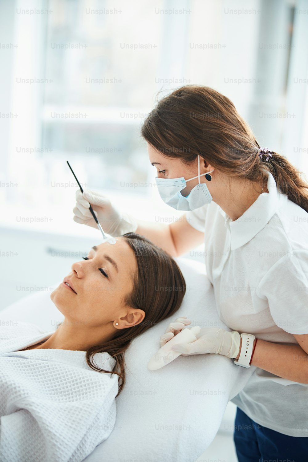 Esteticista focada em luvas nitrílicas espalhando um produto de cuidados da pele sobre a testa da paciente jovem do sexo feminino