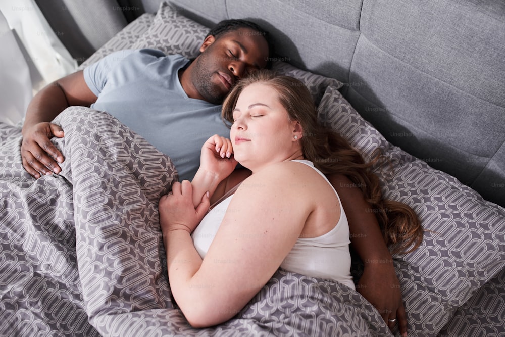 Love and tenderness. Image of young loving multiracial couple laying at the bed indoors at home and hugging while sleeping. Husband and wife enjoying of free time with each other. Stock photo