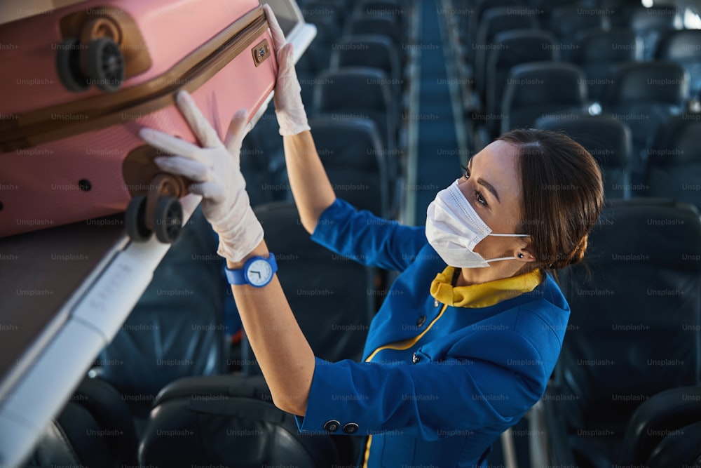 Female cabin attendant wearing safety mask and raising her arms for pushing wheeled suitcase inside overhead bin
