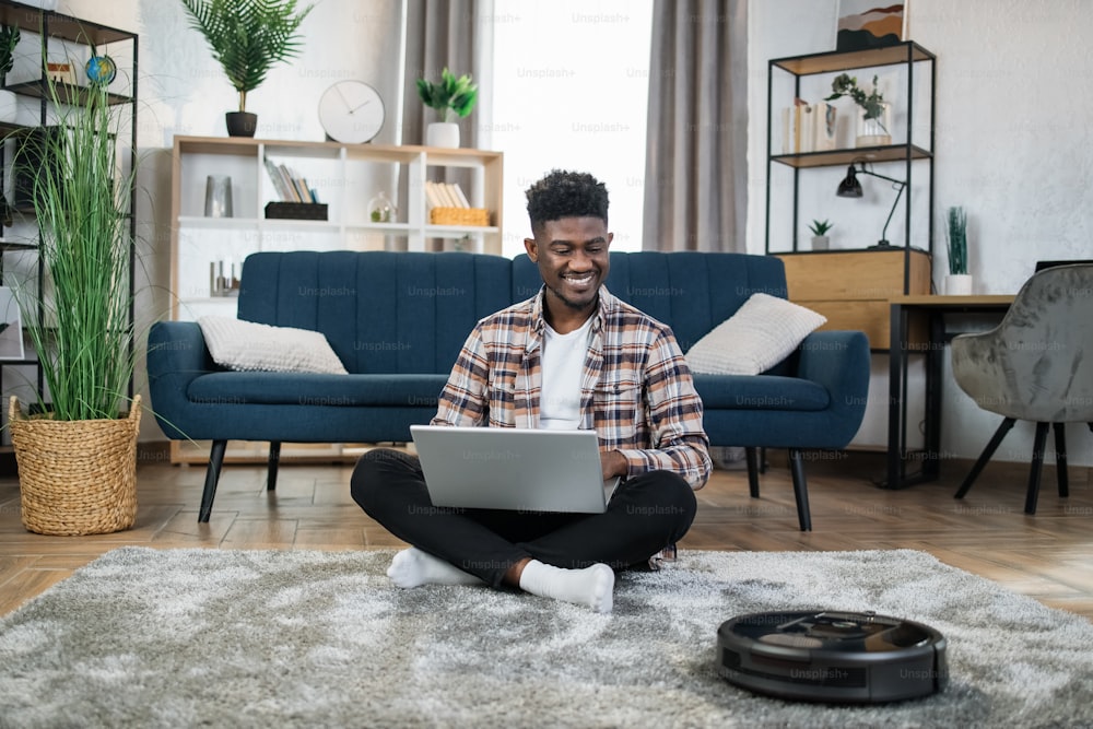 Happy afro-american man sitting on floor with laptop on knees and enjoying how modern vacuum cleaner cleaning his apartment. Household and technology concept.